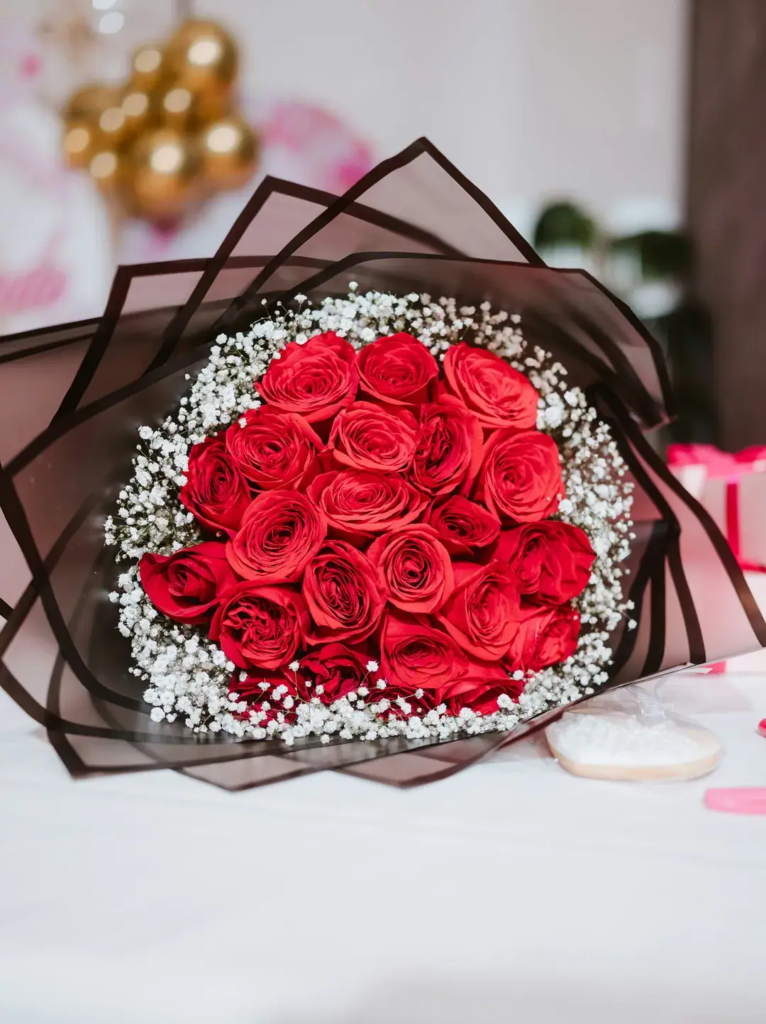 A bouquet of red roses ringed with baby's breath, wrapped in sheer black paper, lying on a white table with gold balloons behind.